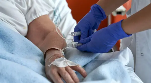 Pielęgniarka w niebieskich rękawiczkach przygotowuje się do usuwania cewnika w domu pacjenta, podłączając strzykawkę do linii infuzyjnej.