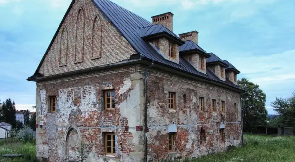 Zniszczony ceglany budynek z drewnianymi oknami i ciemnym dachem, otoczony zielenią.