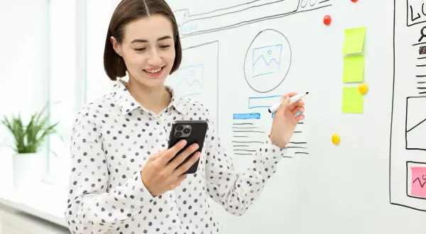 Kobieta z telefonem i markerem przy tablicy z projektami stron. Wyjaśnia, że tekst wyjustowany co to znaczy, pokazując przykłady.
