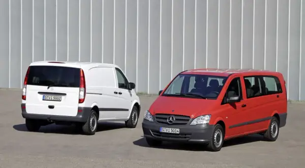 Dwa busy Mercedes-Benz: biały towarowy i czerwony osobowy, stojące na tle szarej ściany.