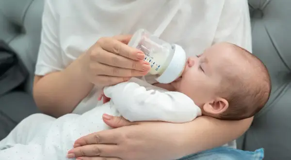 Une main tient un biberon pour nourrir un bébé endormi. La question de savoir si peut on donner de l'eau du robinet à un bébé est importante.