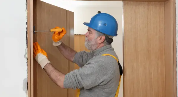 Hombre con casco azul y guantes naranjas ajusta una puerta de contrachapado con un destornillador.