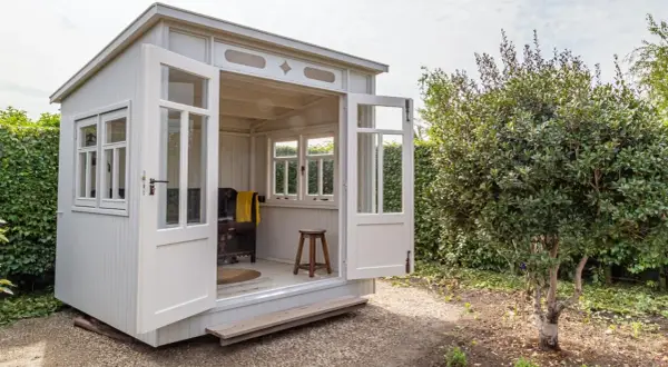 Eine charmante Gartenlaube mit offenen Türen, bereit, mit gemütlichen Möbeln eingerichtet zu werden. Ein Hocker und eine Decke deuten auf eine entspannte Atmosphäre hin.