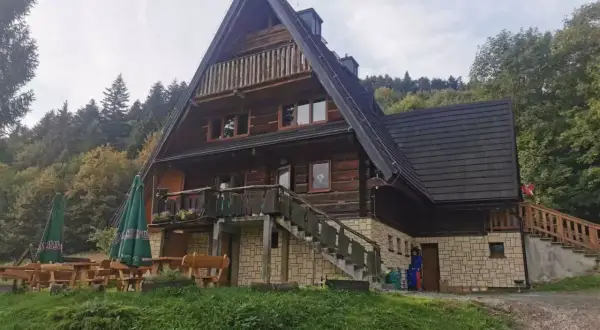 Drewniana bacówka z tarasem i schodami, otoczona zielenią i lasem.
