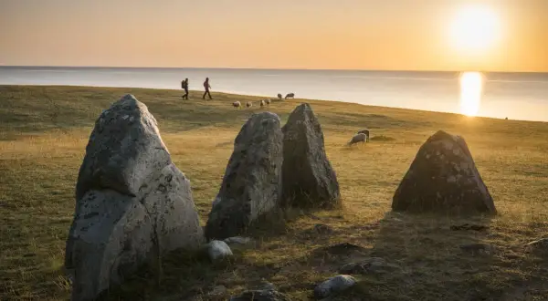 Stora stenar på en äng vid havet under en gyllene solnedgång i Skåne. Får betar och två personer promenerar i fjärran.