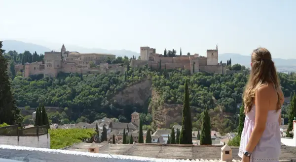 Una mujer contempla la Alhambra, un lugar imprescindible que ver en Granada. El palacio se alza majestuoso sobre un monte verde.