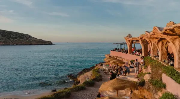 Menschen genießen den Sonnenuntergang am Strand von Ibiza, in der Nähe eines einzigartigen ashram-ähnlichen Gebäudes am Meer.