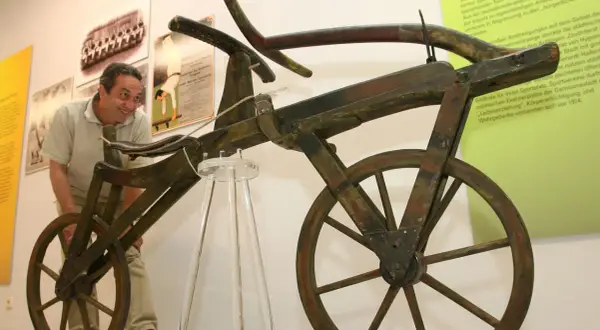 Ein Mann betrachtet das erste Fahrrad der Welt, ein hölzernes Gefährt ohne Pedale, das an ein Laufrad erinnert.