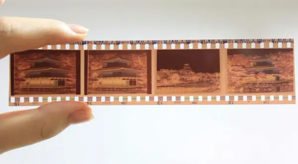 En hand håller upp en filmremsa med fyra bilder från en analog kamera. Bilderna visar en vacker byggnad med japansk arkitektur, omgiven av höstfärger.