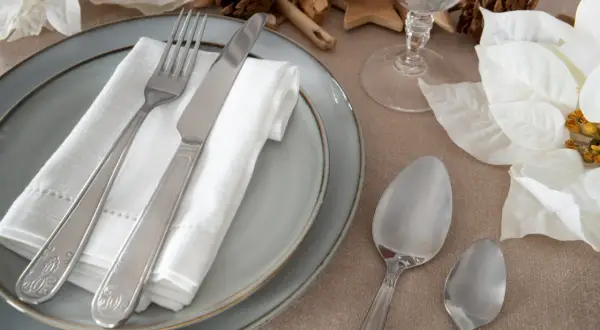 Elegancko ułożone sztućce na talerzu, widelec po lewej, nóż po prawej. Obok łyżki i kieliszek.