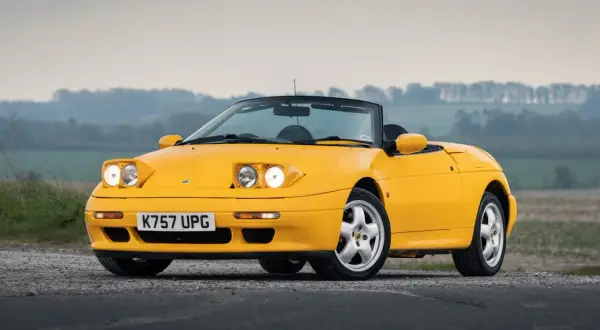 Gul Lotus Elan M100 cabriolet parkerad på grusväg med dimmig bakgrund.