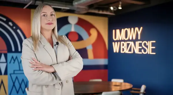 Kobieta w beżowej marynarce z mikrofonem, na tle neonu "UMOWY W BIZNESIE". Czy vider jest bezpieczny?