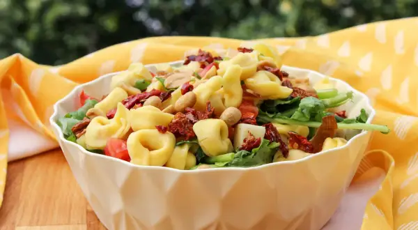 Ein köstlicher tortellini salat mediterran mit sonnengetrockneten Tomaten und Nüssen, serviert in einer weißen Schüssel auf einem Holztisch.