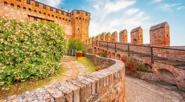 Un sentiero in mattoni conduce al **belvedere ostrense**, con un muro di rose bianche e la rocca medievale sullo sfondo.