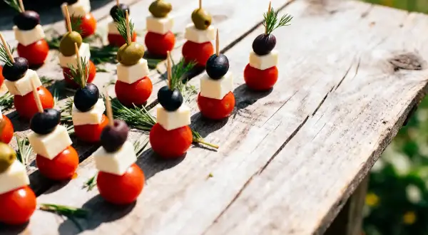 Des mini brochettes apéritives sans cuisson : tomates cerises, dés de fromage, olives noires et vertes, garnies de romarin frais sur une table en bois.