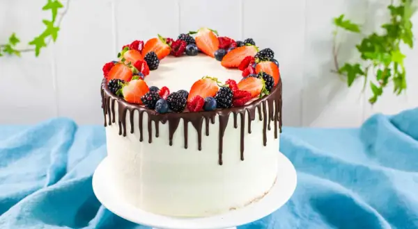 Torta bianca con glassa al cioccolato che cola, decorata con fragole, mirtilli e lamponi freschi. Un drip cake perfetto per ogni occasione.