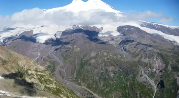 Elbrus, najwyższy szczyt Kaukazu, w chmurach, z lodowcami i zielonymi dolinami poniżej.