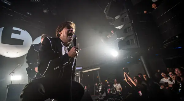 The Hives setlist is being performed by a charismatic frontman in a black suit, belting into a microphone on a smoky stage. The crowd is visible, enjoying the energetic show.