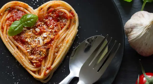 Herzförmige Pasta-Gerichte mit Tomatensoße, Basilikum und Parmesan. Daneben liegen ein Löffel, eine Gabel, Knoblauch und Tomaten.