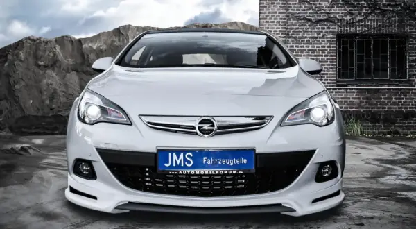 Biały Opel Astra J GTC z tuningiem JMS, z tablicą rejestracyjną "JMS Fahrzeugteile", na tle kamieniołomu i ceglanego budynku.