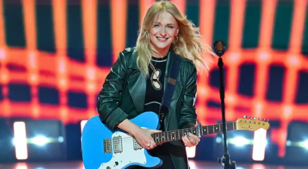 Uśmiechnięta polska piosenkarka z blond włosami gra na niebieskiej gitarze elektrycznej na tle pomarańczowych świateł.