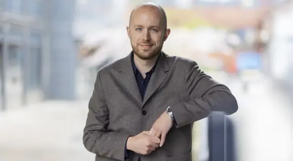 Viktor Barth Kron, en man med kortklippt hår och skägg, bär en brun kavaj och en mörkblå skjorta. Han lutar sig mot ett räcke utomhus.