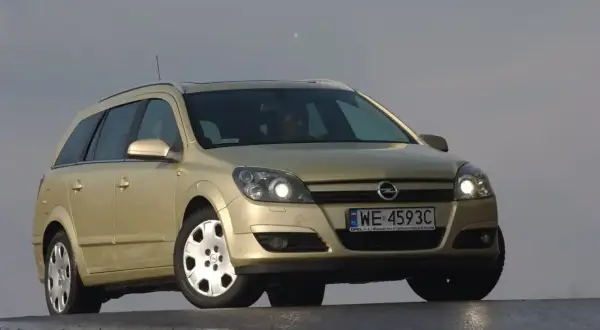 Złoty Opel Astra H kombi z włączonymi światłami. Przód samochodu z wyraźnie widocznymi żarówkami.