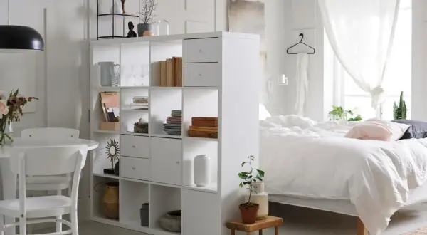 Ein heller Raum mit einem weißen Regal, das als Trenner dient. Links ein Essbereich, rechts ein Bett – ein cleverer wohn und schlafzimmer in einem raum.