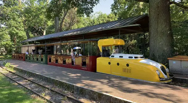 Die gelbe Lokomotive der Schlossbahn Karlsruhe steht bereit. Fahrgäste sitzen in den roten und grünen Waggons unter einem schattigen Dach.