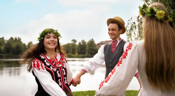 Trzy osoby w tradycyjnych strojach ludowych trzymają się za ręce, tańcząc na tle jeziora.