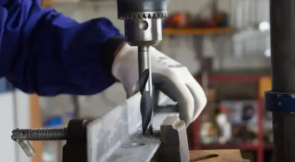 Ręka w rękawicy obsługuje wiertarkę stołową, wiercąc w metalowym profilu. Widać części do wiertarki stołowej i wióry metalowe.