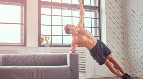 Mężczyzna ćwiczy crossfit w domu, wykonując deskę boczną na kanapie. Pokazuje, jak ćwiczyć crossfit w domu bez sprzętu.