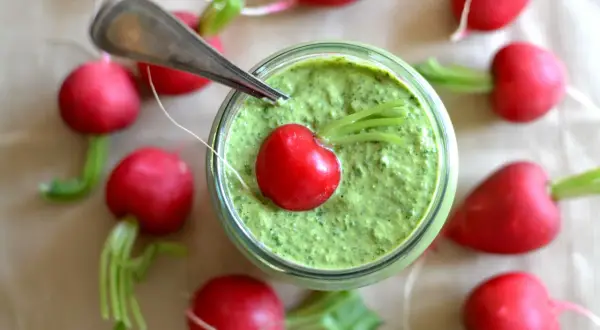 Słoik z zielonym pesto z liści rzodkiewki, ozdobiony jednym rzodkiewkowym serduszkiem. Wokół rozłożone są świeże rzodkiewki.