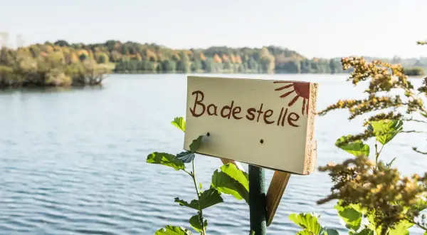Schild "Badestelle" am See mit Bäumen im Hintergrund.