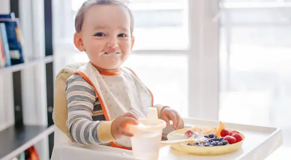 Kleiner Junge mit Heidelbeeren und Joghurt im Gesicht, der an seinem Becher nippt.