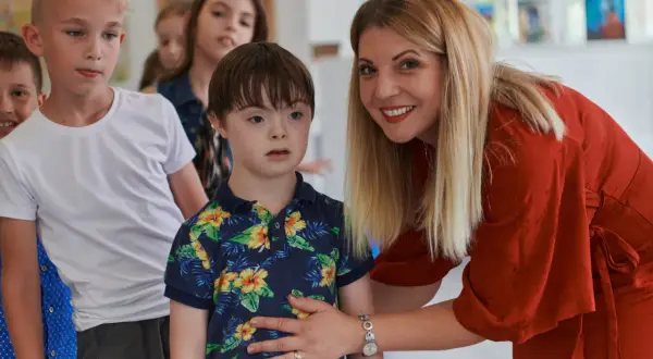 Eine Frau in Rot lächelt und legt ihre Hand auf die Schulter eines Jungen mit Down-Syndrom. Man darf behindert sagen, wenn man über Inklusion spricht.