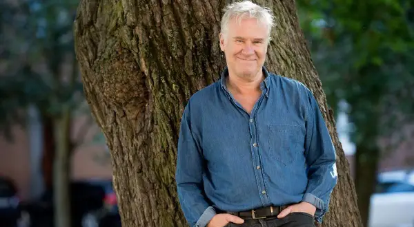 Jacob Nordenson lutar sig mot ett träd, klädd i jeansskjorta och mörka jeans, med ett leende.
