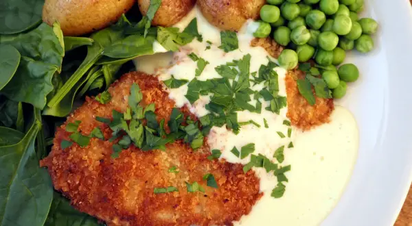 Krispig schnitzel med krämig sås till schnitzel, serverad med små potatisar, gröna ärtor och färsk spenat.