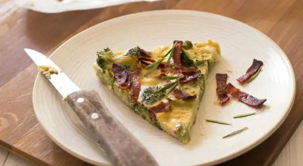 Une part de quiche sans pâte aux brocolis et lardons, garnie de romarin, sur une assiette avec un couteau.