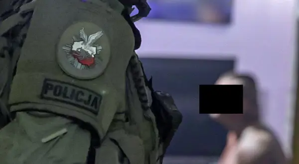 Funkcjonariusz CBŚ w kamizelce z napisem POLICJA i godłem Polski. W tle mężczyzna z zamaskowaną twarzą.