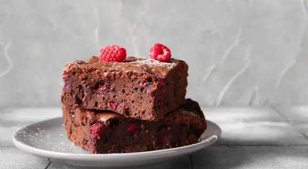 Dwa wilgotne brownie z malinami, posypane cukrem pudrem, na szarym talerzyku. Idealne na deser.