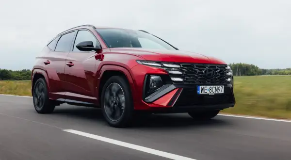Czerwony Hyundai Tucson 1.6 T-GDI N Line pędzi po drodze, otoczony zielonymi polami i drzewami.