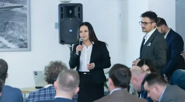 Kobieta w czarnej marynarce przemawia do grupy ludzi, trzymając mikrofon.