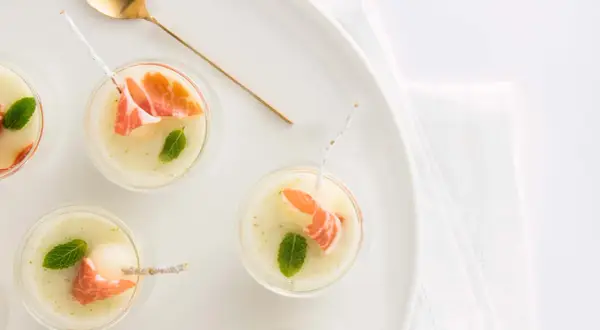 Delicioso chupito de melón con jamón serrano y menta, servido en copas pequeñas con una cuchara dorada.