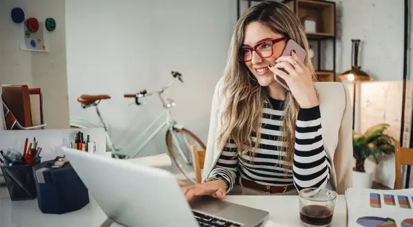 Frau am Telefon, lächelnd, am Laptop arbeitend. Hinter ihr ein Fahrrad. Perfekt für den **iu semesterbeginn**-Stress.