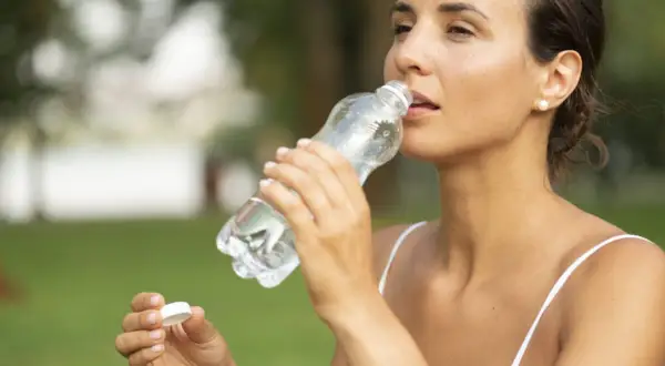 Kobieta pije wodę z plastikowej butelki na tle zieleni.