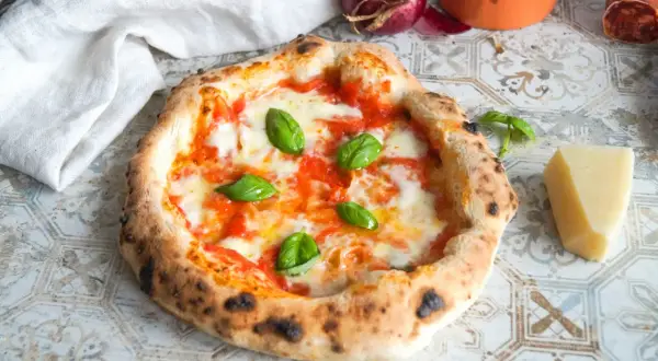 Eine authentische Pizza Napoletana mit frischem Basilikum, geschmolzenem Mozzarella und Tomatensauce. Daneben ein Stück Parmesan und eine rote Zwiebel.