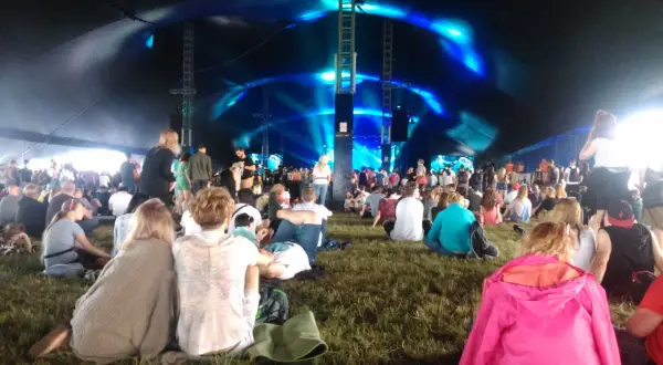 Widownia siedzi na trawie pod namiotem, czekając na koncert. Niebieskie światła oświetlają scenę, tworząc atmosferę festiwalu Open'er.