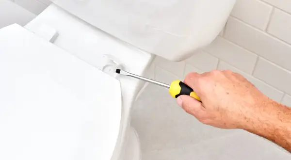 A hand uses a screwdriver to tighten a bolt, demonstrating how to install a toilet seat.