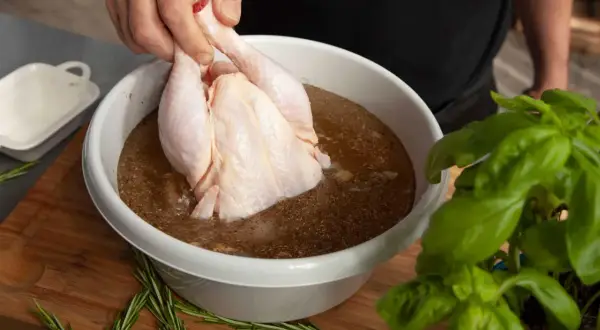 Ein rohes Hähnchen wird in eine Schüssel mit einer würzigen Flüssigkeit getaucht, um Fleisch in Salzlake einzulegen. Rosmarin und Basilikum liegen daneben.